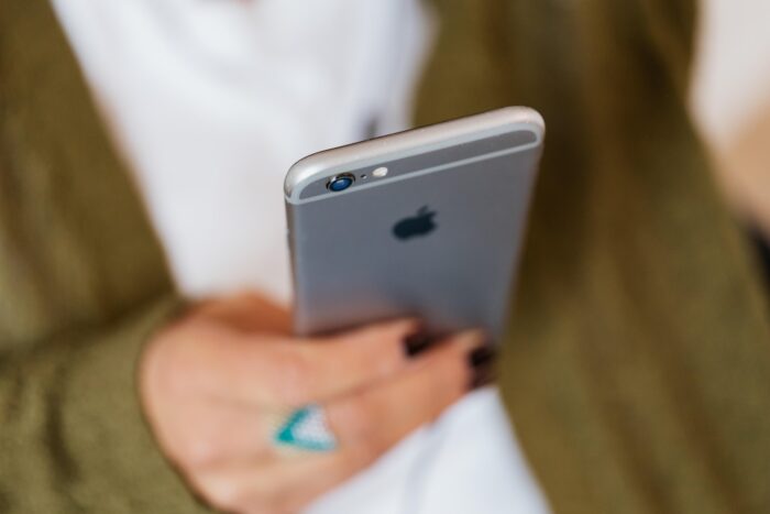 Blurred woman wearing a long sleeved green sweater with a white shirt underneath it holding onto an Apple cell phone. The womans nails are painted a dark brown and the right had has a turquoise ring on her middle finger. 