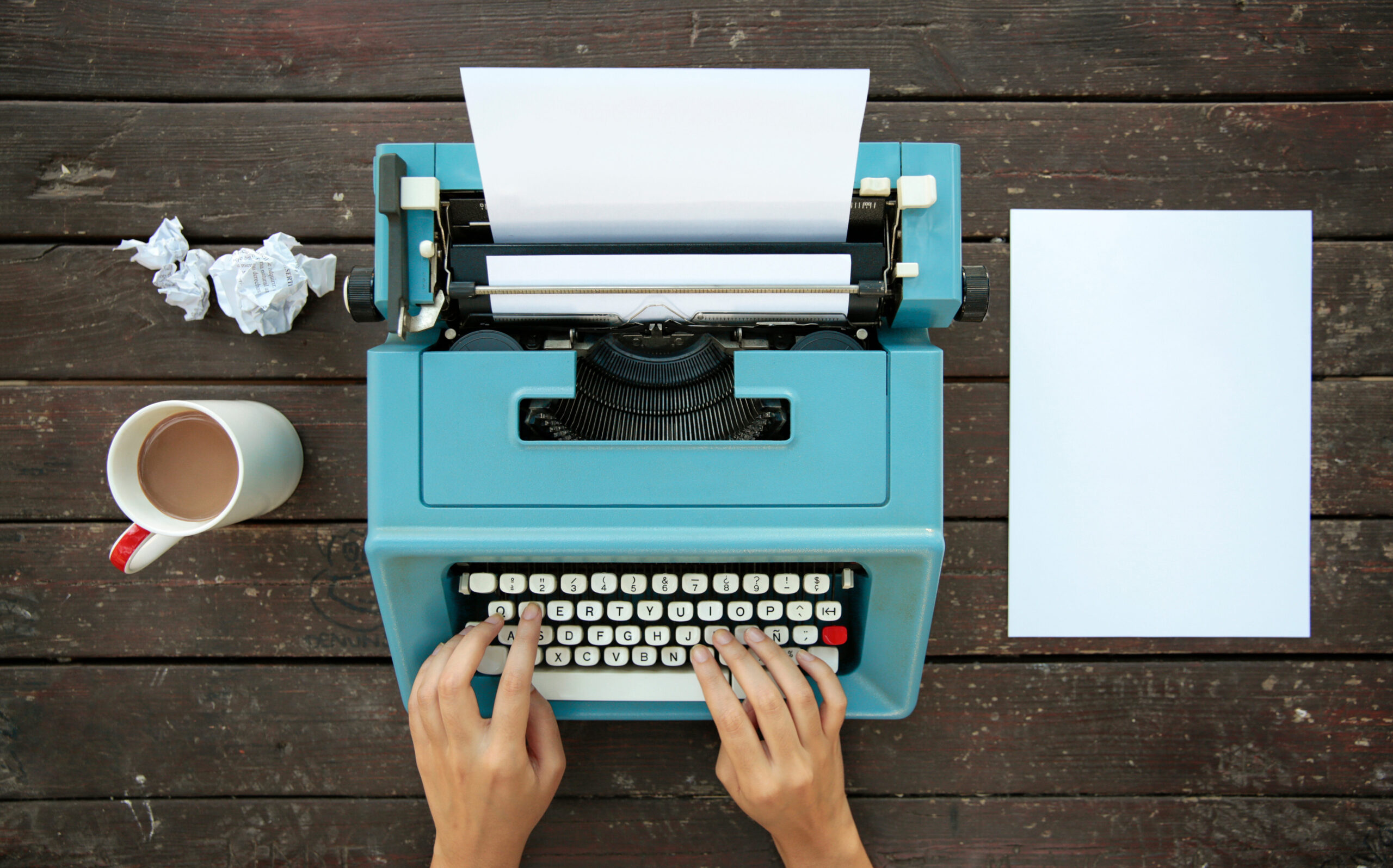 Hands are typing on a turquoise colored type-writer that is sitting on a wooden plank table with balled up pieces of papers off to the side, right behind a cup of coffee. A blank piece of paper is laying on the table to the right of the type-writer.