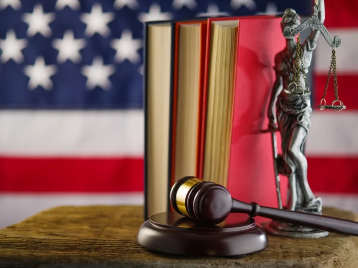 A wooden desk with 3 books being held up by Lady Justice, a dark mahogany wood gavel with a brass ring placed in front with an American flag  blurred in the background.